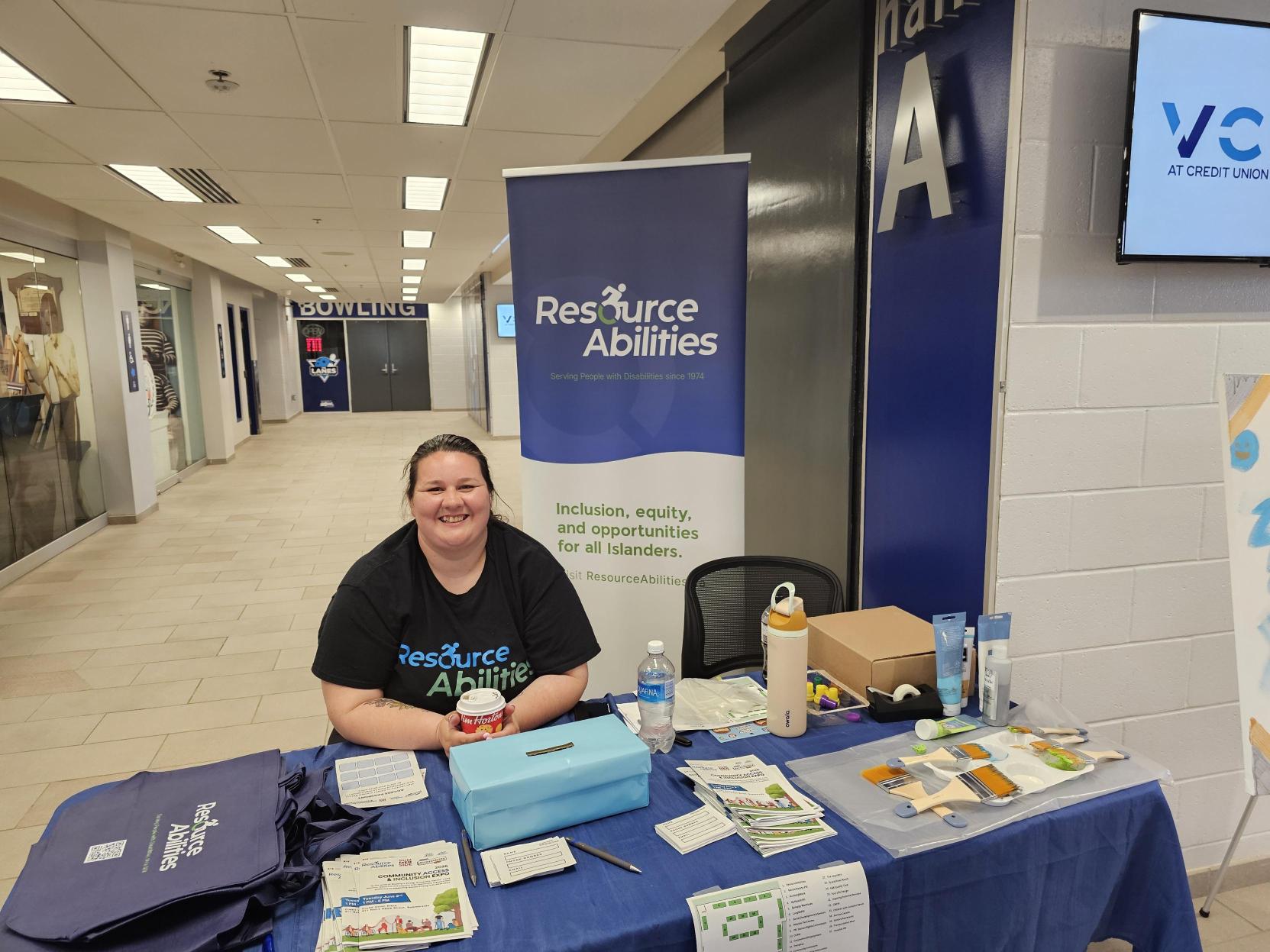A woman in a black ResourceAbilities tshirt sits behind a booth covered in materials and swag promoting Resourceabilities. Behind her stands a tall ResourceAbilities banner.